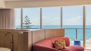 Une pièce moderne avec un canapé rouge aux lignes courbes, une grande fenêtre donnant sur la mer, un plancher en bois et une salle de bain au style minimaliste.