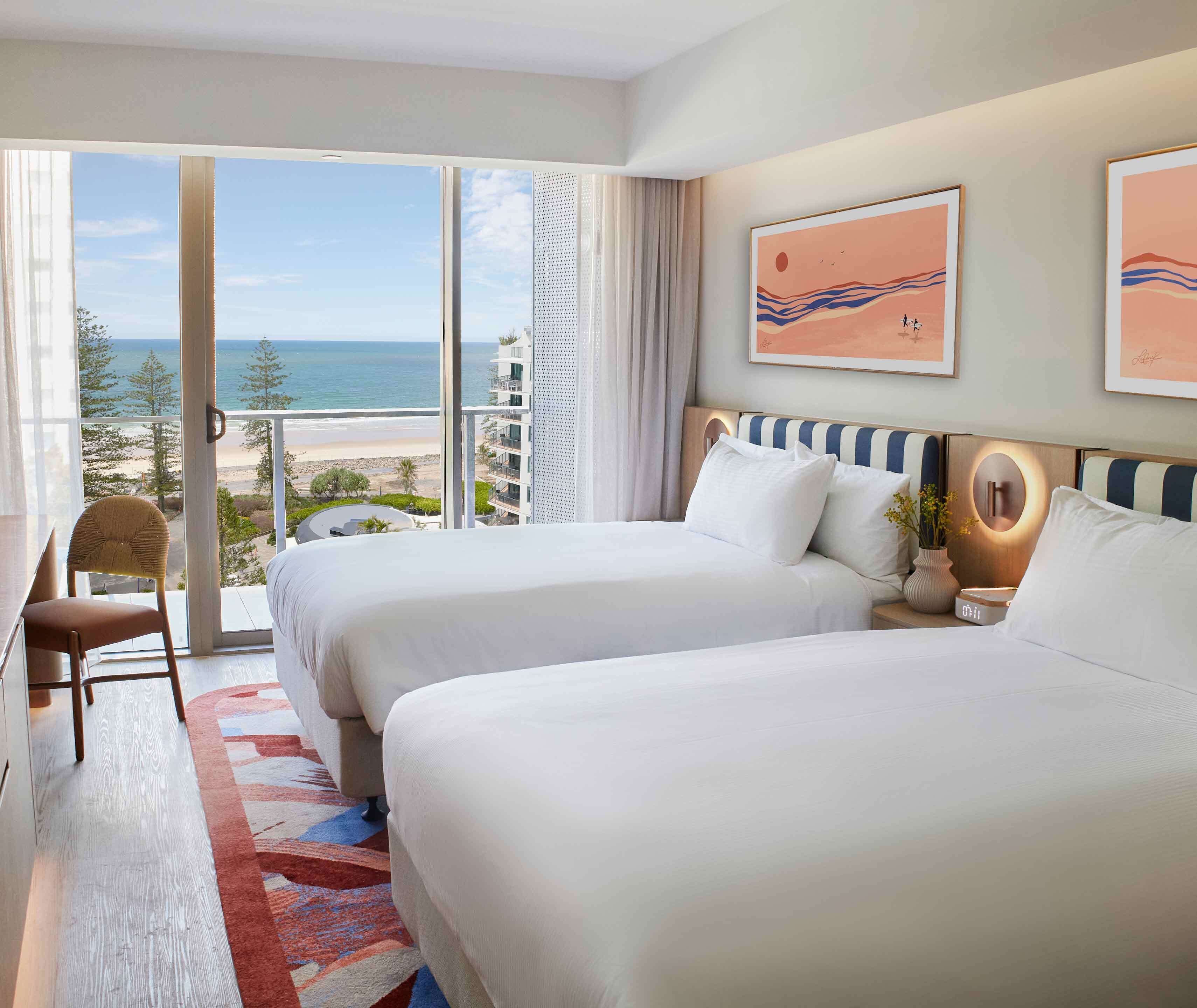 Hotelkamer met twee bedden, een bureau, een stoel en een groot raam met uitzicht op het strand en de oceaan.
