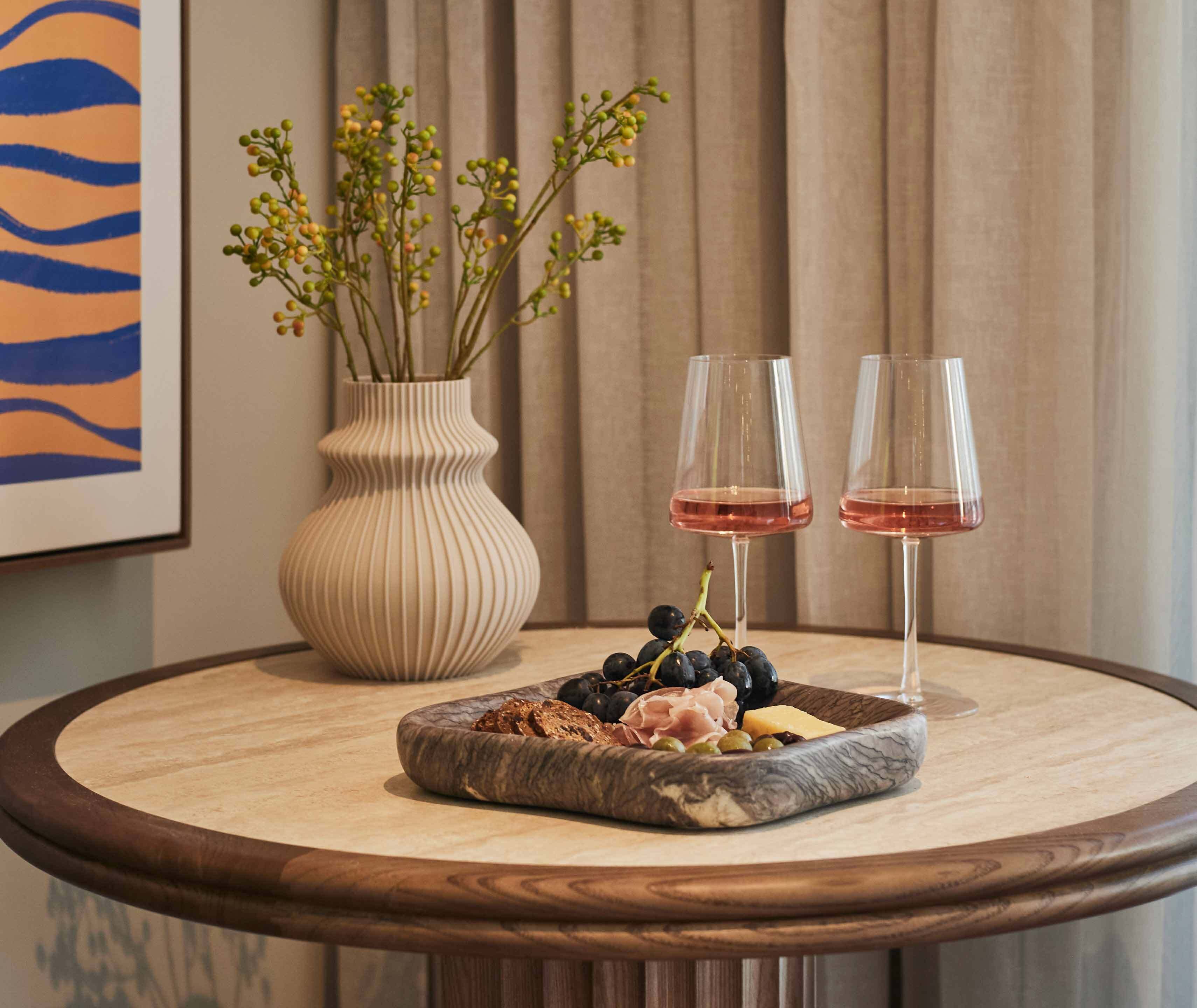 A round wooden table with a marble top, a vase with green foliage, two wine glasses, and a charcuterie board with various meats, cheeses, and grapes.
