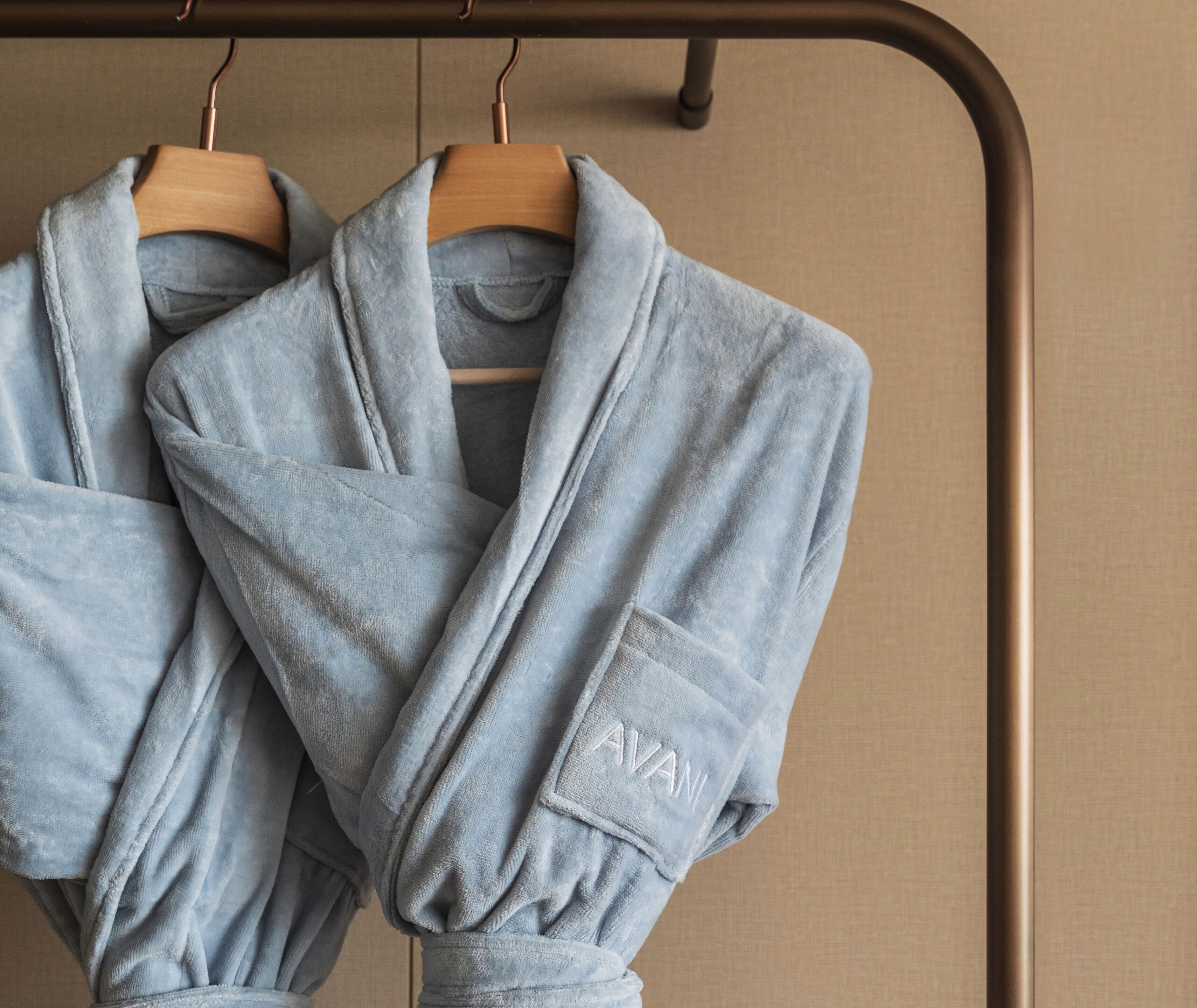Two light blue robes hanging on a wooden hanger against a beige wall.