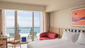 A hotel room with a large bed, a view of the ocean, a dining area with a table and chairs, and a painting on the wall.