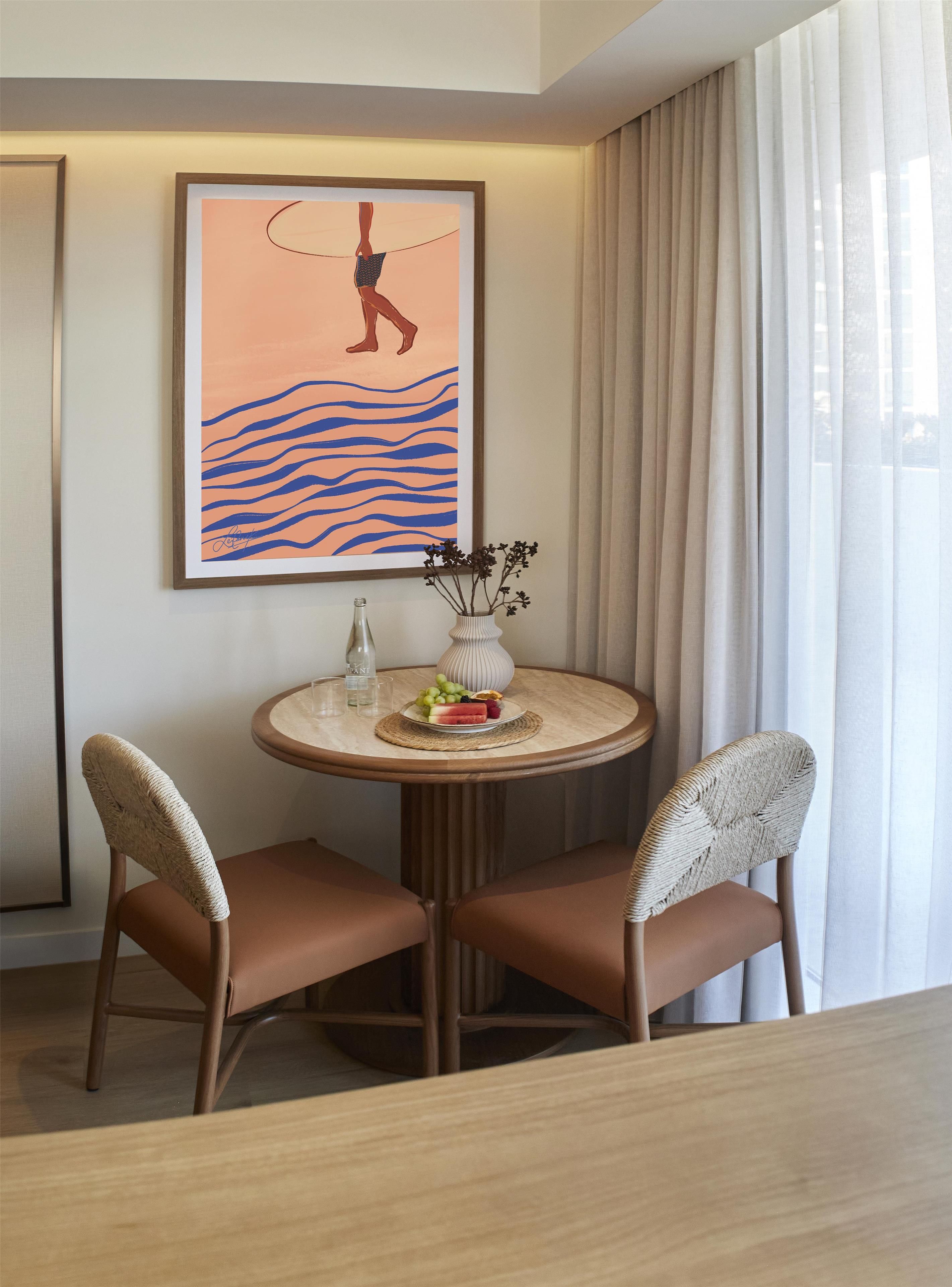 A hotel room with a round table, two chairs, a framed artwork, a vase with flowers, and a bottle.