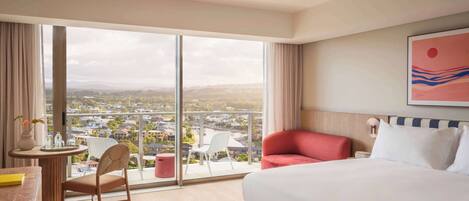 A hotel room with a large window, a bed, a chair, a small table, and a painting on the wall.