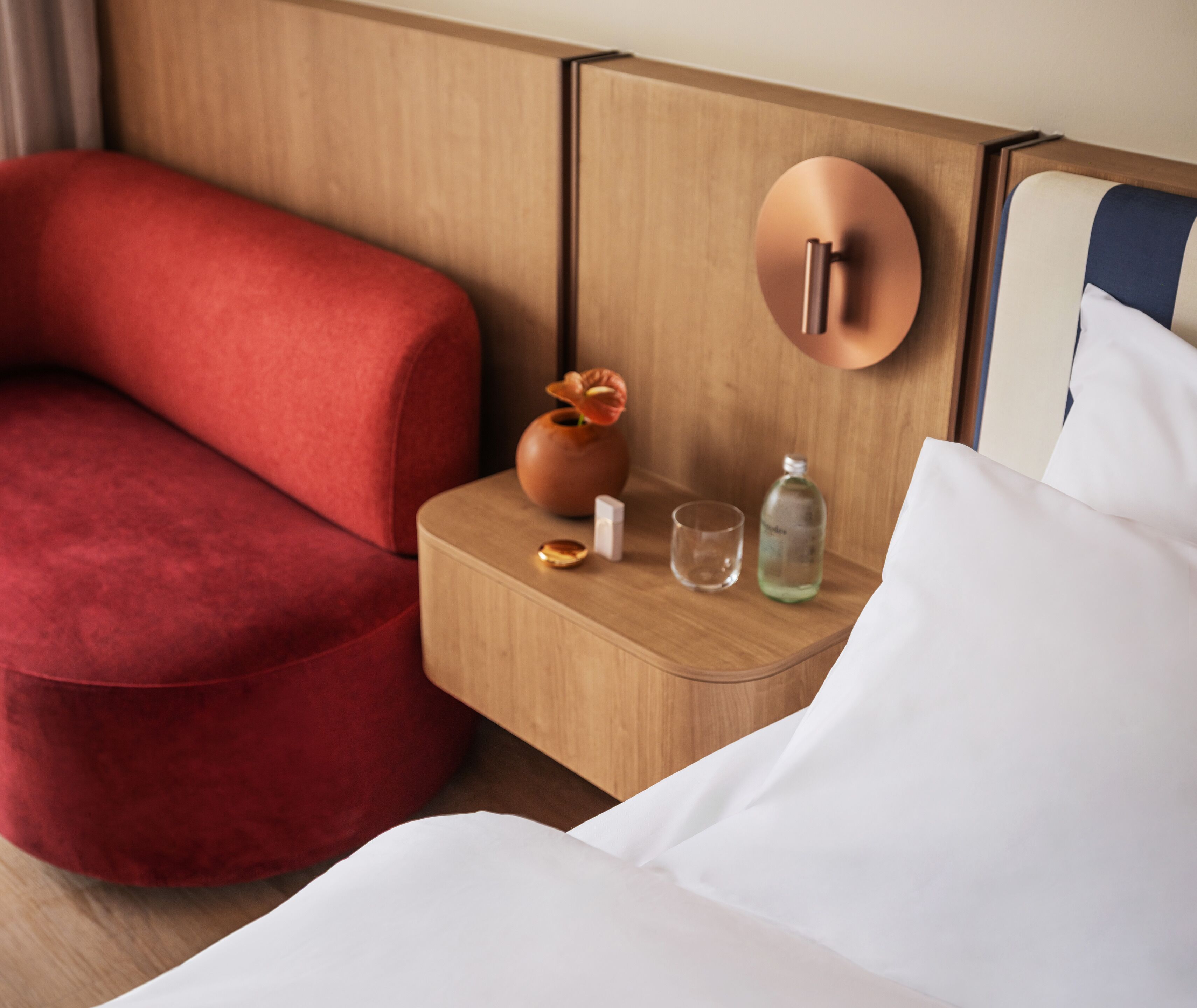 Une chambre d’hôtel avec un fauteuil à revêtement rouge, une table de chevet en bois ornée d’un vase et d’une bouteille, et un lit avec du linge de lit blanc.