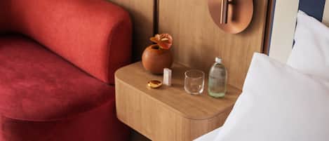 Una habitación de hotel con una silla tapizada de rojo, una mesita de noche de madera con un florero y una botella, y una cama con ropa de cama blanca.
