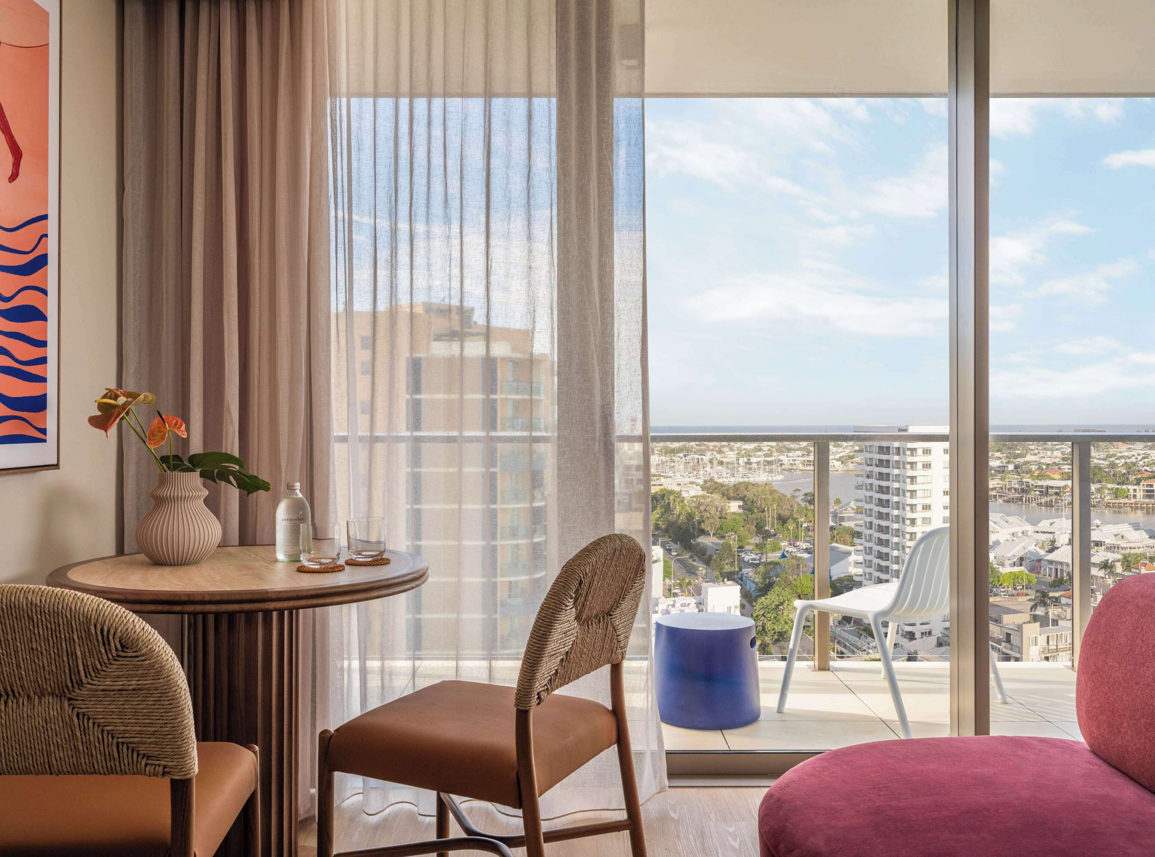A modern hotel room with a balcony, a round table with a vase of flowers, a chair, and a view of the cityscape.