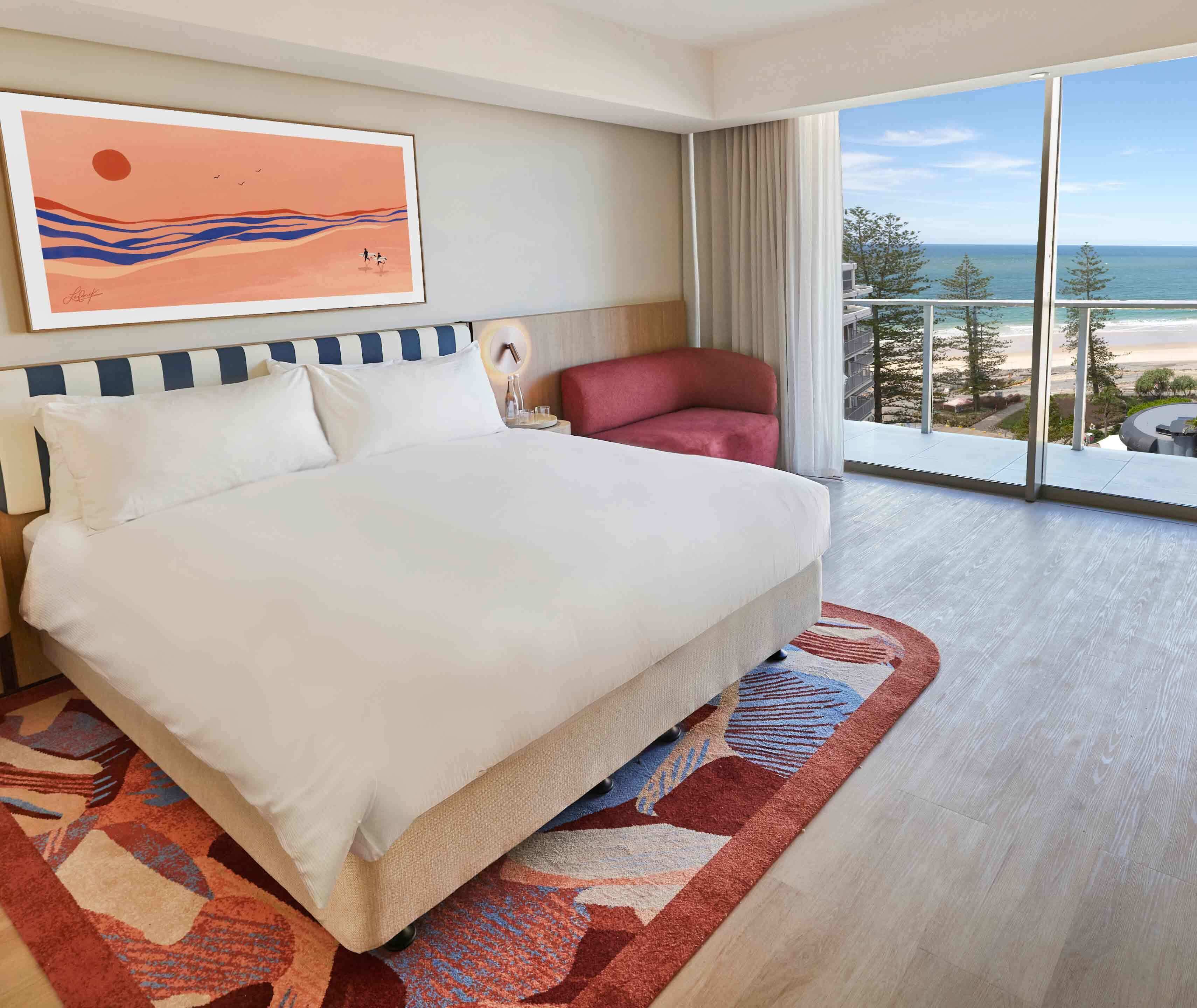Een hotelkamer met een groot bed, een rode stoel, een schilderij en een balkon met uitzicht op de oceaan.