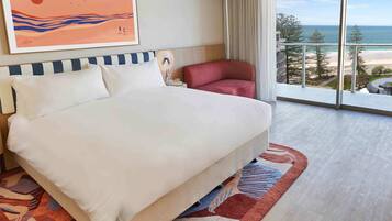 A hotel room with a large bed, a red chair, a painting, and a balcony with a view of the ocean.