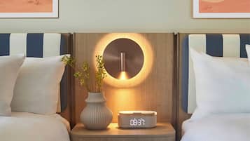 A hotel room with two beds, a bedside table with a clock, a vase with flowers, and framed artwork on the wall.