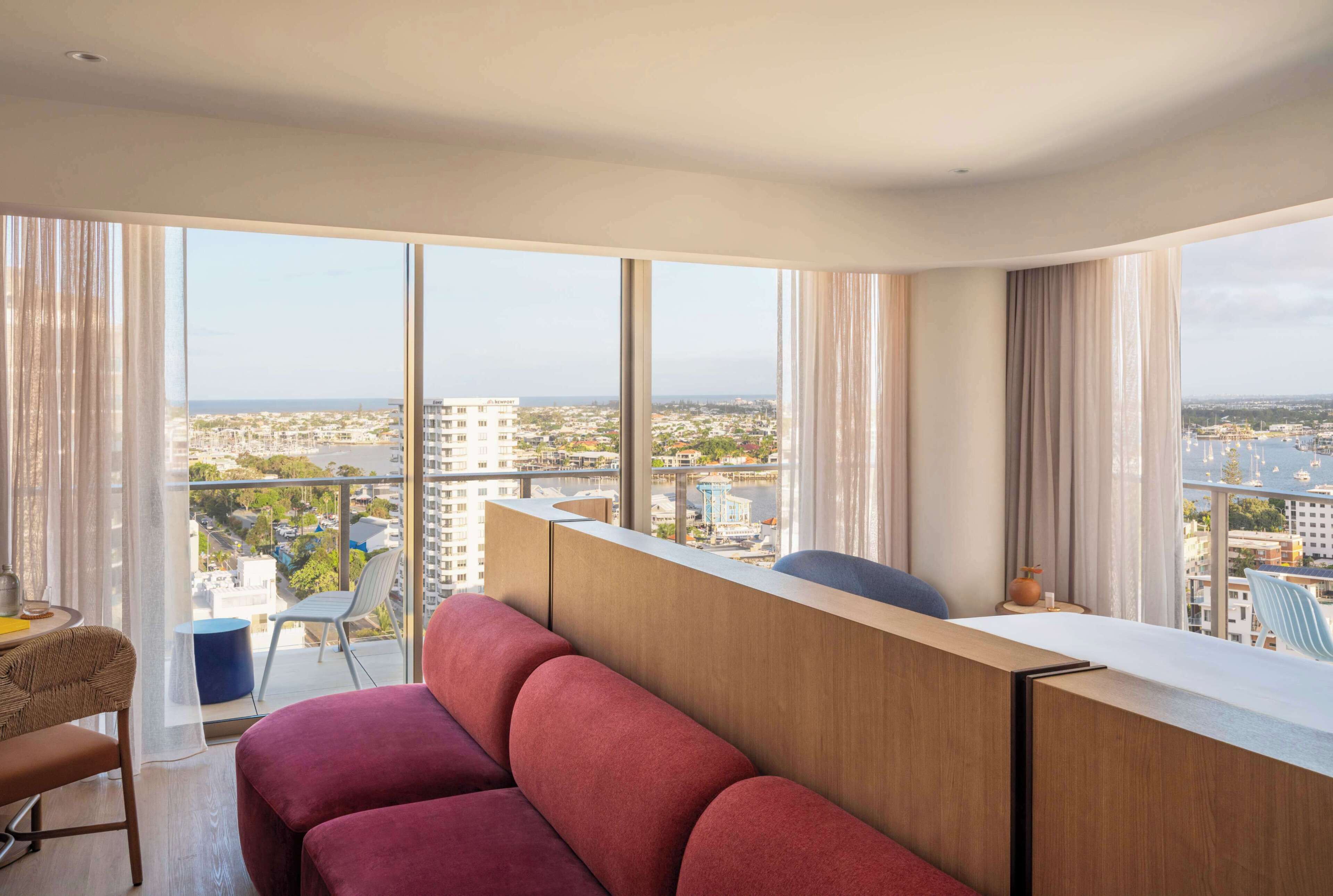 Ein modernes Hotelzimmer mit großem Balkon, einem roten Sofa, einem kleinen Tisch und Blick auf die Stadt.