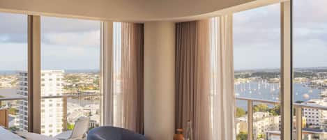A hotel room with a large bed, a chair, a small table with a vase and a bottle, and a view of a cityscape and marina through large windows.