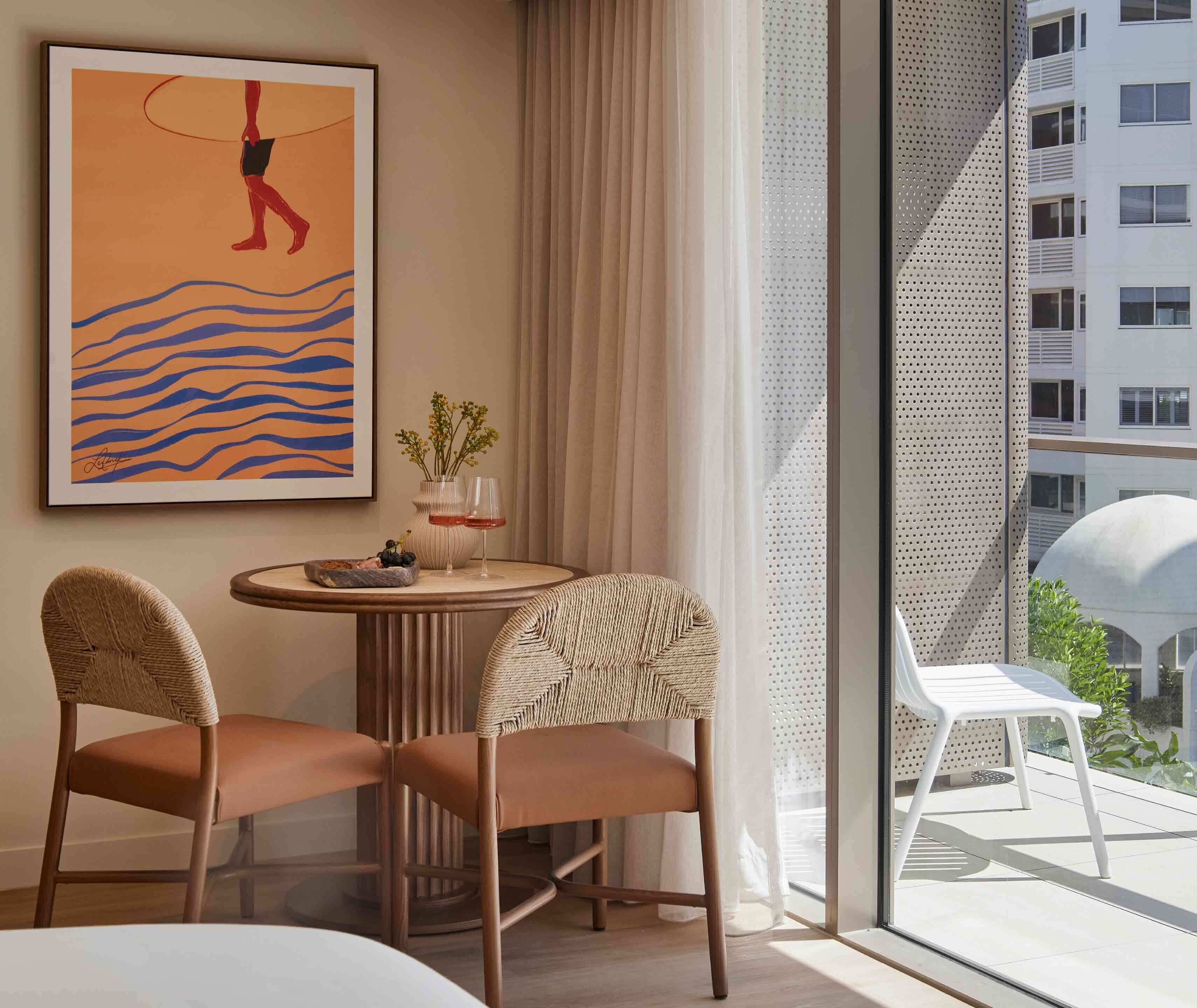 A hotel room with a large window, a round table, two wicker chairs, a framed artwork, and a view of a building and greenery outside.