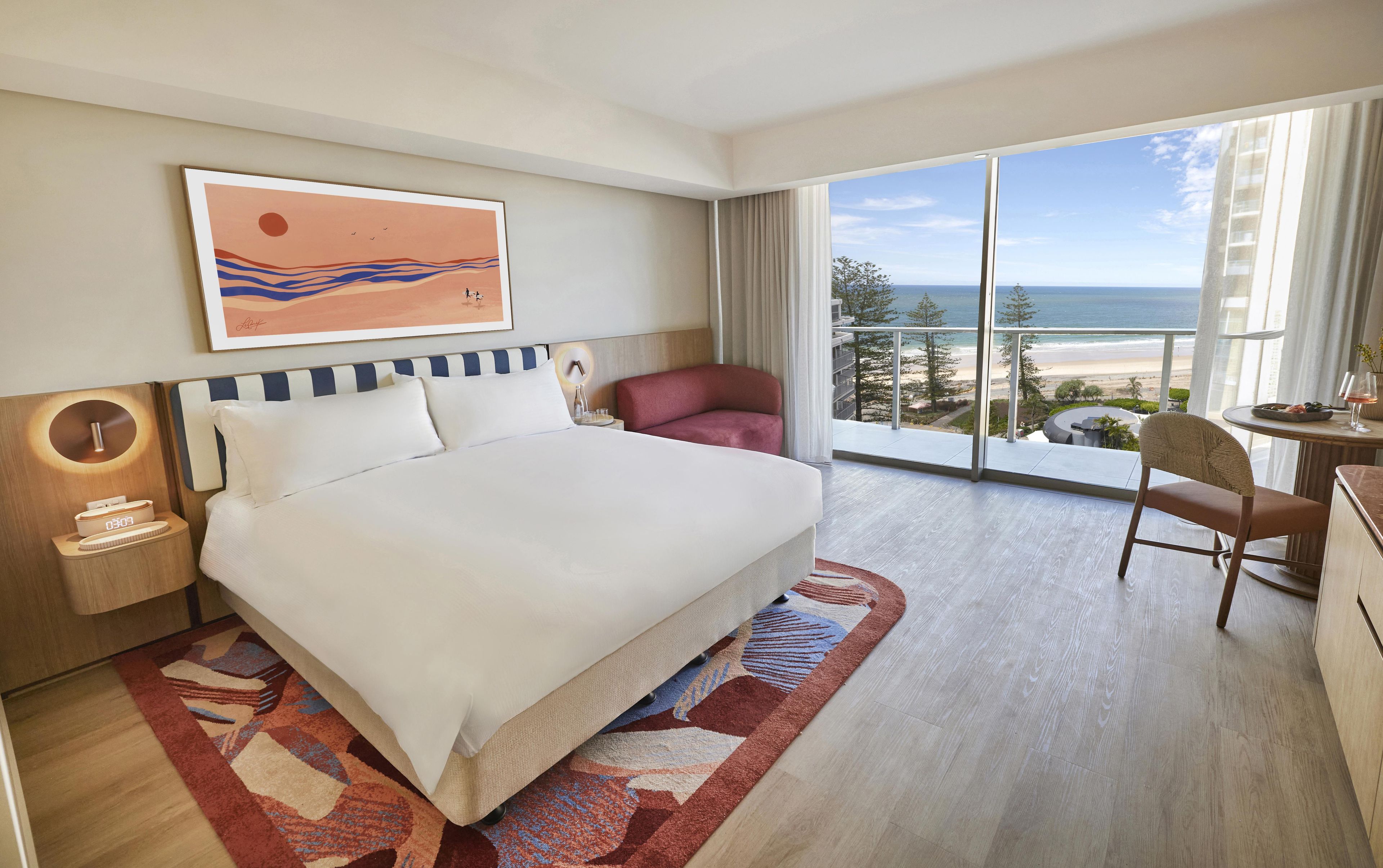 A hotel room with a large bed, a desk, a chair, a view of the beach, and a painting on the wall.