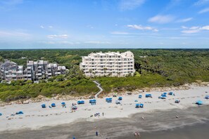 A beachfront hotel with multiple balconies, a sandy beach with umbrellas, and a forested area in the background.
