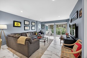 A modern living room with a grey sofa, a coffee table, and a TV. There are framed pictures on the wall and a large window with a view of greenery.
