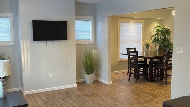 Una sala de estar con piso de madera, un televisor de pantalla plana montado en la pared, un comedor con mesa y sillas, y una planta grande en maceta.