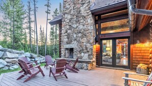Fachada de piedra con chimenea, terraza de madera, sillas Adirondack y vista a árboles.