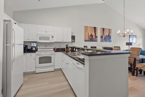 A modern kitchen with white cabinets, a built-in oven, a microwave, and a central island with a sink. There are two paintings on the wall and a chandelier above the dining area. - Ocean Keyes Lakeview Condo 2 Blocks From Beach! (North Myrtle Beach)
