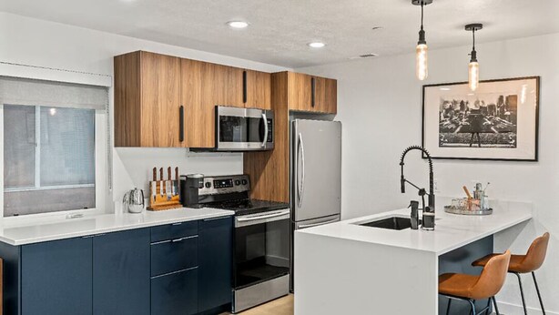 A modern kitchen with dark blue cabinets, a stainless steel refrigerator, a microwave, and an oven. There is a small island with a sink and two bar stools. - SLC downtown hub (Salt Lake City)