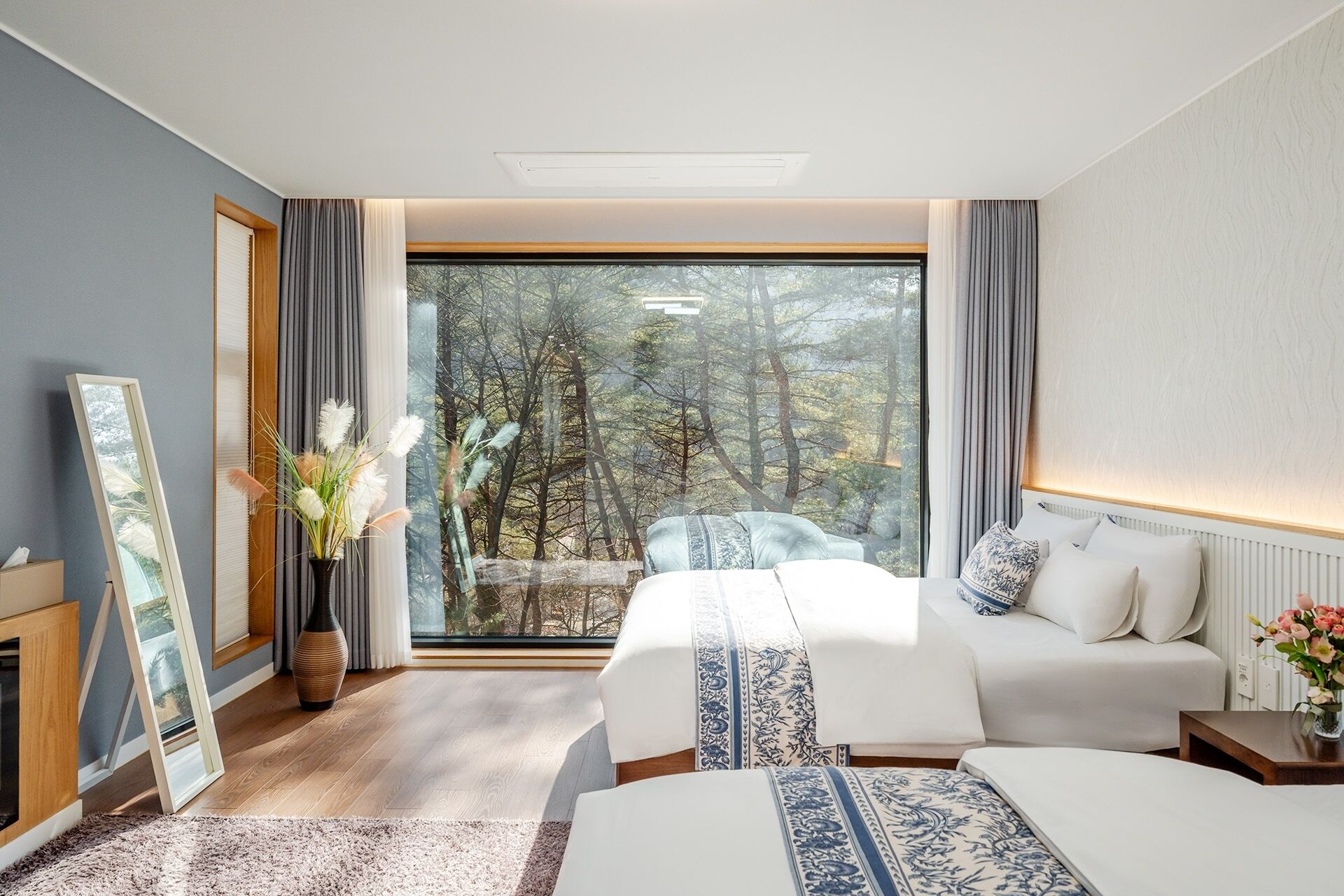 A bedroom with a large window, two beds, a bedside table, and a mirror.