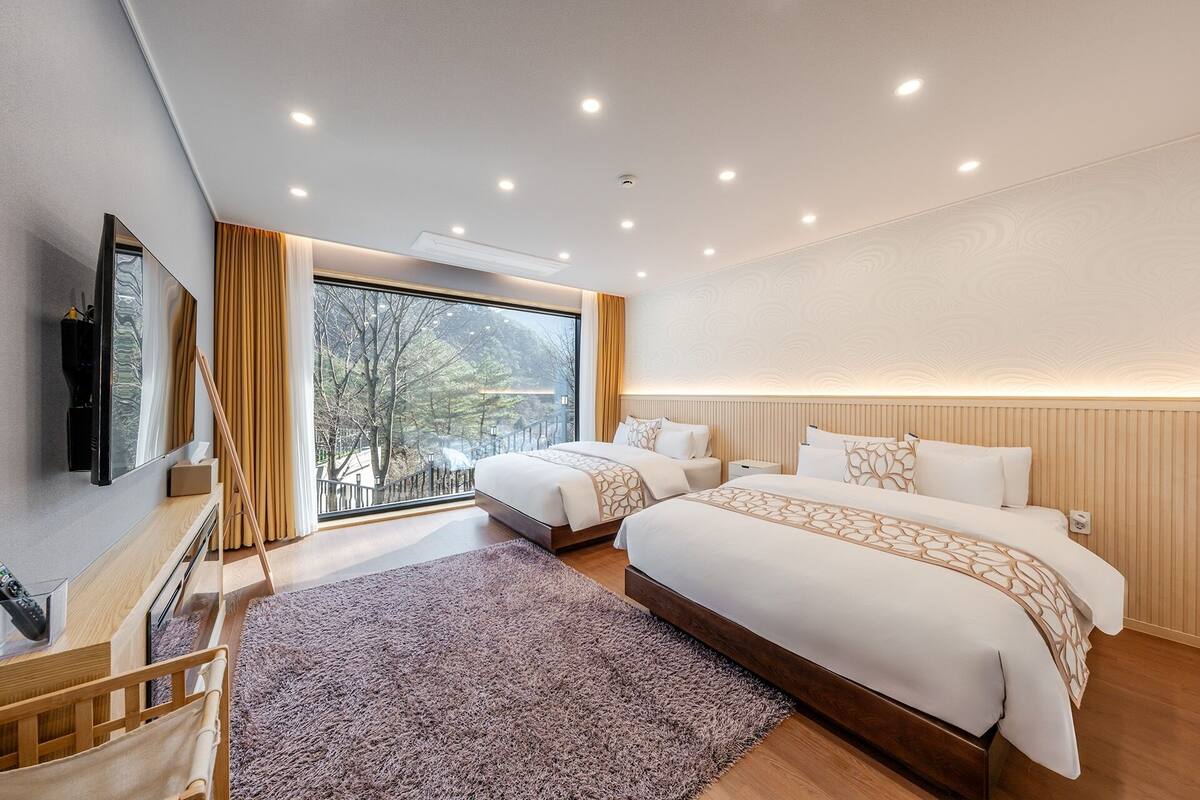 A hotel room with two beds, a large window, a flat-screen TV, and a wooden floor.