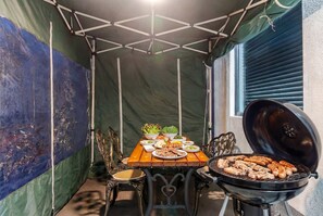 Ett takförsett utomhusområde med plats för måltider, utrustat med en grill, ett bord, stolar och en tältliknande konstruktion.
