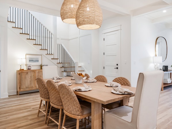 A dining area with a wooden table, wicker chairs, a staircase, and a white door. - 🦩Book Me Baby! Daydream Believer in Seagrove Beach 🦩 (Seagrove Beach)