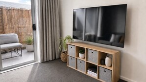 A modern living room with a large flat-screen TV mounted on the wall, a grey sofa, a small wooden console table with storage, and a sliding glass door leading to an outdoor area.