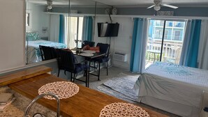 Une pièce comprenant un lit, une table à manger avec des chaises, un téléviseur fixé au mur et un balcon donnant sur un immeuble.