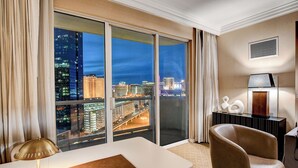 Ein Hotelzimmer mit einer großen Schiebetür, einem Schreibtisch mit Lampe, einem Sessel und Blick auf die nächtliche Skyline.