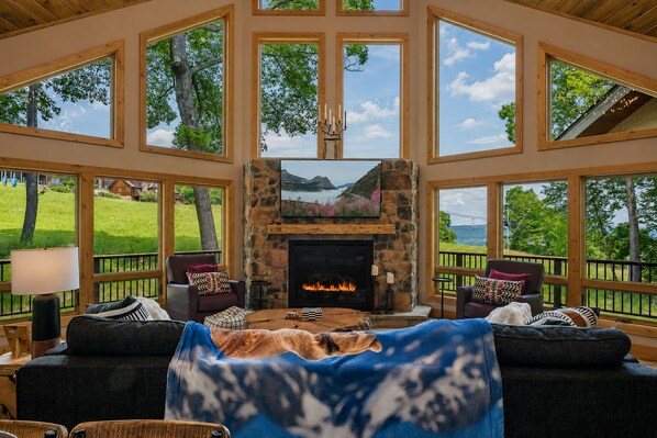 A spacious room with a stone fireplace, large windows, and a view of the outdoors. - Designer Home: Hot Tub Sauna Game Room EV Charger (McHenry)