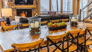 A cozy living room with a stone fireplace, a television mounted above it, and a dining area with wooden furniture and candles. - Designer Home: Hot Tub Sauna Game Room EV Charger (McHenry)