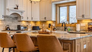 A kitchen with white cabinets, a large island with chairs, a stove, and a window with a view. - Designer Home: Hot Tub Sauna Game Room EV Charger (McHenry)