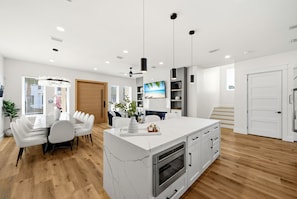 A modern kitchen with a central island, a dining area with a white table and chairs, and a staircase leading to another level. - 5BR, 4.5BA Coastal Escape w/ Pool, Elevator, Near Beach & Attractions (Panama City Beach)
