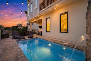 A swimming pool with lounge chairs and string lights, surrounded by a stone wall and a building with large windows. - 5BR, 4.5BA Coastal Escape w/ Pool, Elevator, Near Beach & Attractions (Panama City Beach)