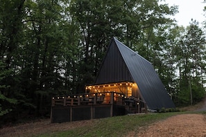 A modern, triangular-shaped building with a deck and string lights, surrounded by dense forest.
