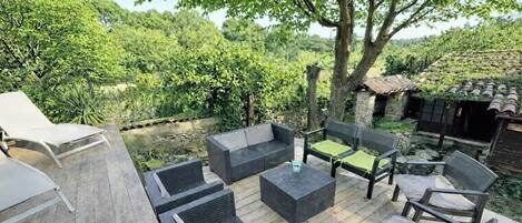 Een terras buiten met rieten meubels, een tafeltje en uitzicht op een tuin en bomen.