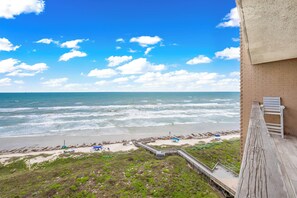 Blick auf einen Strand von einem Balkon mit Holzdeck, einer Ziegelwand und blauem Himmel.