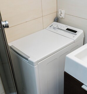 A front view of a compact, modern washing machine in a bathroom with tiled walls and a visible electrical outlet. - Głogowska Comfort Apartment (Poznań)