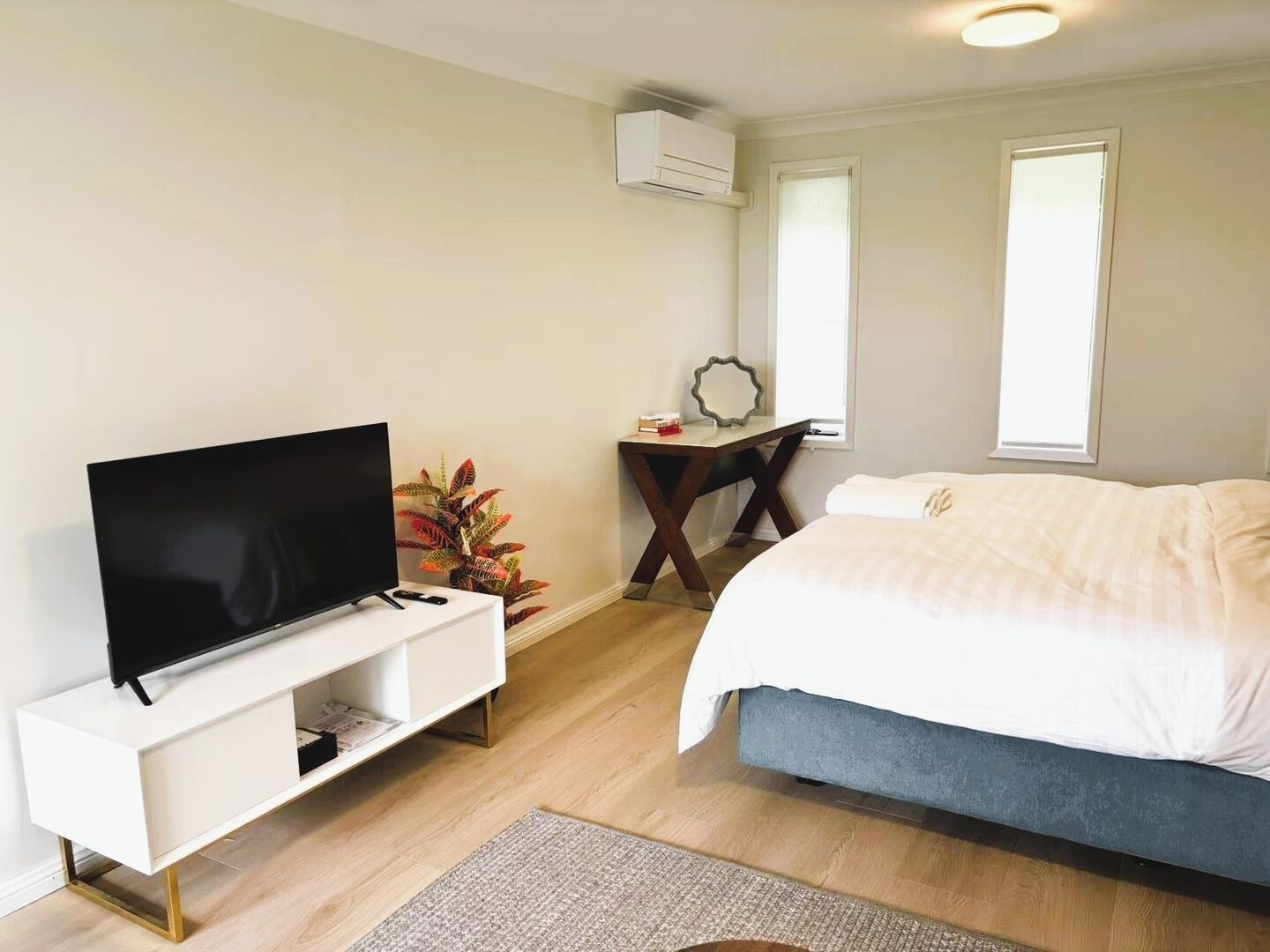 A modern bedroom with a flat-screen TV, a bedside table with a mirror, a bed with white bedding, and a potted plant.