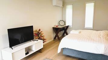 A modern bedroom with a flat-screen TV, a bedside table with a mirror, a bed with white bedding, and a potted plant.