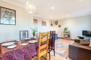 Un área de comedor con una mesa preparada para cuatro personas, un sofá, una estantería y un televisor en una sala de estar.