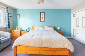Un dormitorio con una cama, dos mesitas de noche, un tocador, un aparador, una ventana con cortinas y una puerta.