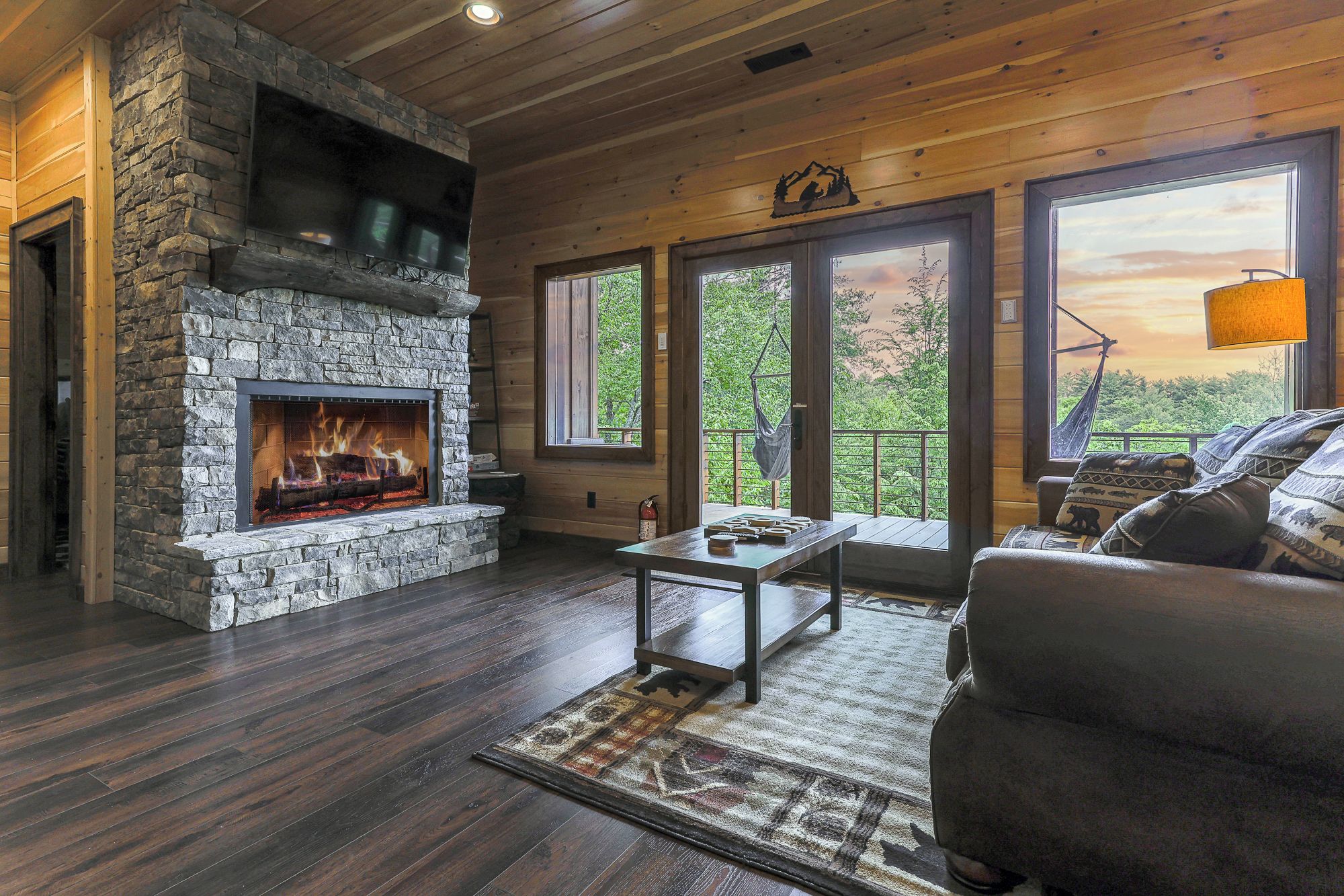 A cozy cabin interior with a stone fireplace, wooden floors, a sofa, and large windows offering a view of the outdoors.