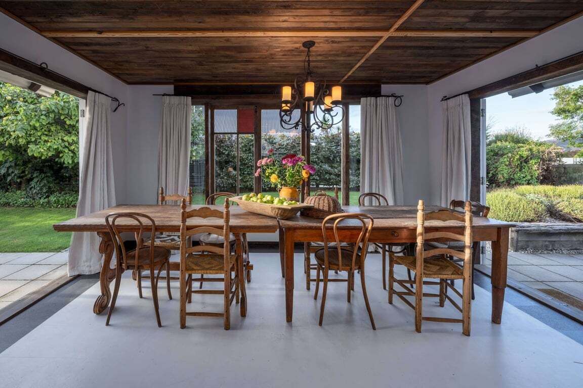 A dining area with a wooden table and chairs, a chandelier, and a view of the garden through large windows.