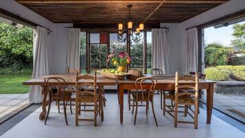 A dining area with a wooden table and chairs, a chandelier, and a view of the garden through large windows.