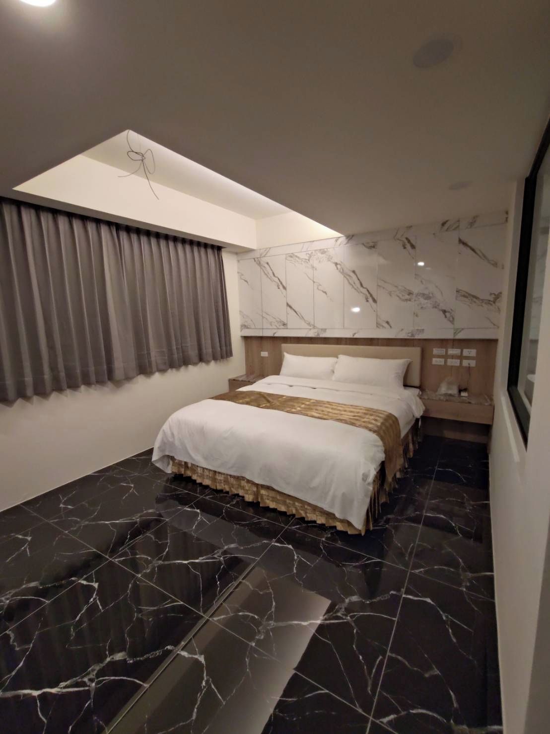 Une chambre d’hôtel moderne avec un lit, une table de chevet, une fenêtre avec des rideaux et un sol en marbre.