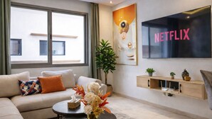 A modern living room with a large window, a sofa, a coffee table, a TV, and a wall-mounted shelf with decorative items. - Maarif 1BR | Garage + Fast WiFi + Netflix (Dar-el-Beida)