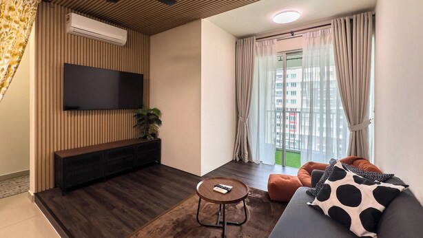 A modern living room with a flat-screen TV mounted on a wooden panel wall, a small round wooden coffee table, a grey sofa with a cow print pillow, and a large window with sheer curtains. - Residensi Suasana Damai by Airhost (Sungai Buloh)