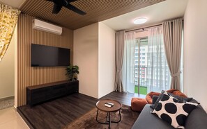 Ein modernes Wohnzimmer mit einem flachbildfernseher an einer Wand aus Holzpaneelen, einem kleinen runden Couchtisch, einem grauen Sofa mit Kissenmuster Kuhfell und einem großen Fenster mit durchscheinenden Vorhängen.